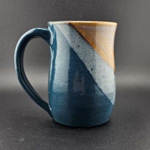May include: A handmade ceramic mug with a large handle. The mug features a color-blocked design with navy blue, white, and brown sections. The mug is suitable for hot or cold beverages.