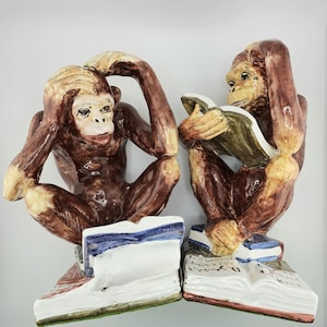 May include: Two ceramic monkey figurines in brown and tan hues. One covers its ears, the other reads a book. Each sits atop a stack of books with blue, white, and red accents. The figurines have a glossy finish.