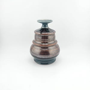 May include: A handcrafted ceramic jar with a lid. The jar has a bulbous base, a cylindrical midsection, and a smaller top with a lid. The colors are a mix of brown, bronze, and teal, with a metallic sheen. The jar is on a white background.