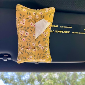 May include: A yellow fabric tissue holder with a floral pattern of white flowers and green leaves. The holder is rectangular with a pocket and is hanging from a car visor. White tissues are partially visible.