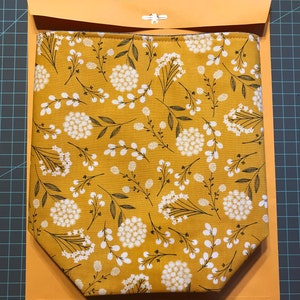 K&ouml;nnte beinhalten: Eine gelbe Stofftasche mit einem wei&szlig;en Blumenmuster. Die Tasche ist gefaltet und in einem braunen Umschlag gelegt. Der Text "mush boho" steht auf dem Umschlag.