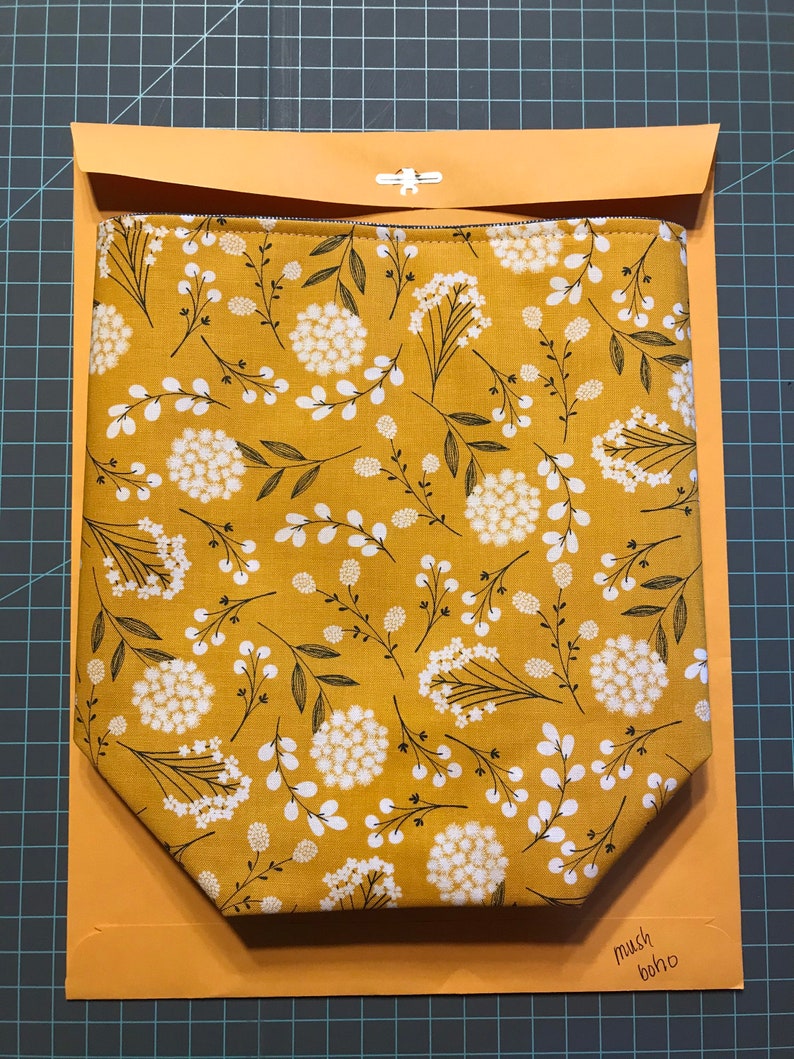 K&ouml;nnte beinhalten: Eine gelbe Stofftasche mit einem wei&szlig;en Blumenmuster. Die Tasche ist gefaltet und in einem braunen Umschlag gelegt. Der Text "mush boho" steht auf dem Umschlag.