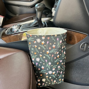 May include: A black fabric car trash can with a floral pattern in shades of pink, orange, and white. The trash can is hanging on the side of a car seat.
