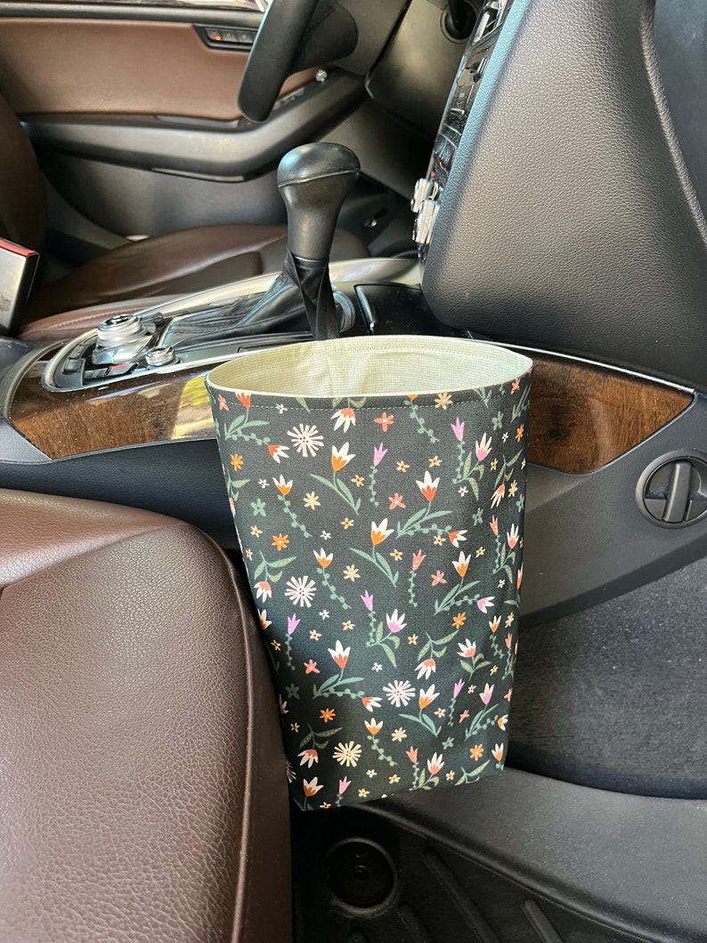 May include: A black fabric car trash can with a floral pattern in shades of pink, orange, and white. The trash can is hanging on the side of a car seat.
