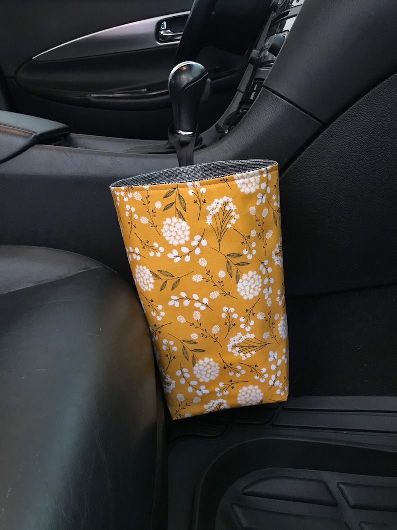 K&ouml;nnte beinhalten: Ein gelber und wei&szlig;er Auto-M&uuml;lleimer mit floralem Muster und einem schwarzen Riemen, der an der Schalthebel in einem Auto h&auml;ngt.