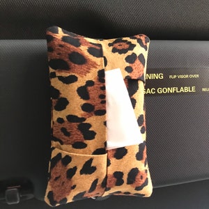 May include: A brown and black leopard print tissue box holder with a white tissue inside. The holder is hanging on the back of a car seat.