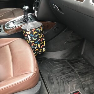 May include: A black car interior with a brown leather seat and a black floor mat with the WeatherTech logo. A fabric trash can with a butterfly pattern is hanging from the side of the seat.