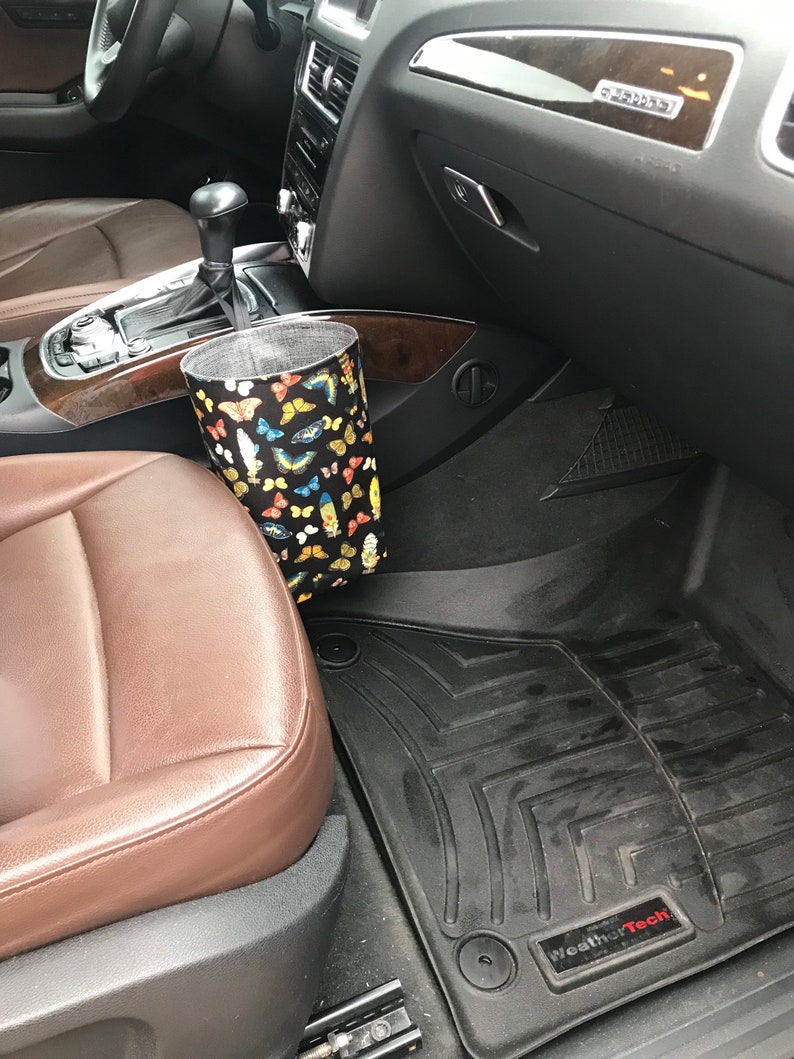 May include: A black car interior with a brown leather seat and a black floor mat with the WeatherTech logo. A fabric trash can with a butterfly pattern is hanging from the side of the seat.