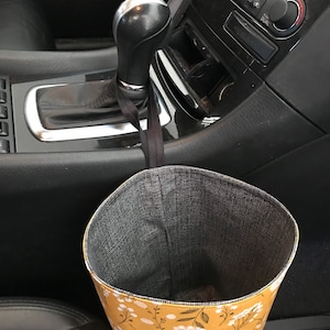 K&ouml;nnte beinhalten: Ein gelber und wei&szlig;er, mit Blumen gemusterter Stoffm&uuml;lleimer, der an der Schaltung eines Autos h&auml;ngt.