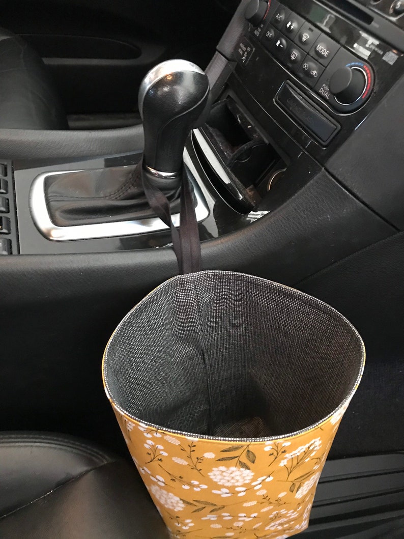 K&ouml;nnte beinhalten: Ein gelber und wei&szlig;er, mit Blumen gemusterter Stoffm&uuml;lleimer, der an der Schaltung eines Autos h&auml;ngt.