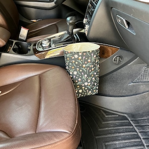 May include: A black and white floral patterned trash can with a beige liner is hanging on the side of a car's center console.
