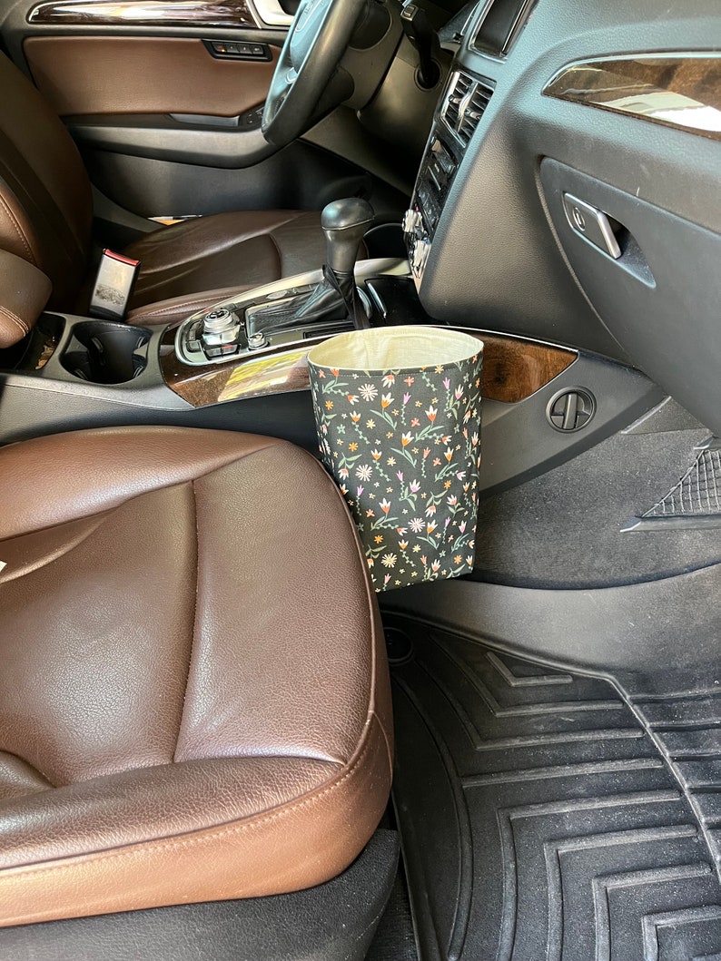 May include: A black and white floral patterned trash can with a beige liner is hanging on the side of a car's center console.