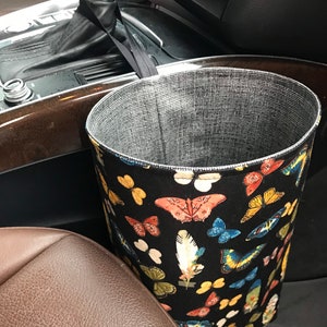May include: A black fabric car trash can with a butterfly and feather print. The trash can is hanging on the side of a car seat.