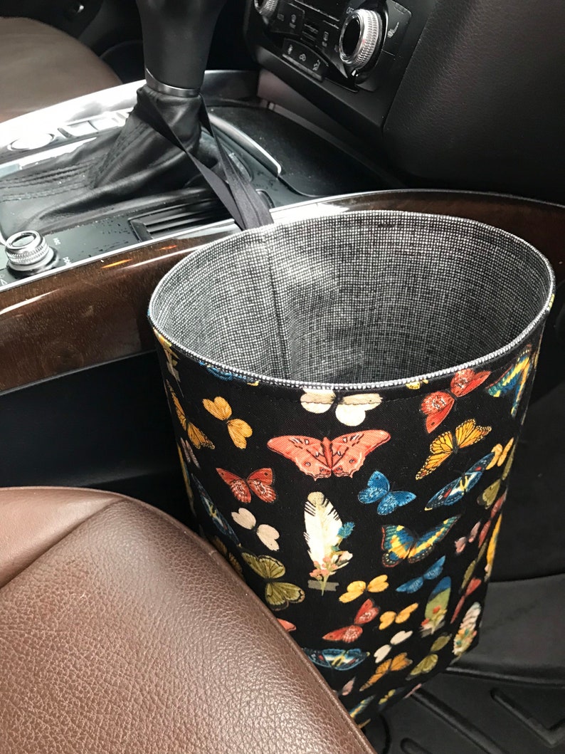 May include: A black fabric car trash can with a butterfly and feather print. The trash can is hanging on the side of a car seat.