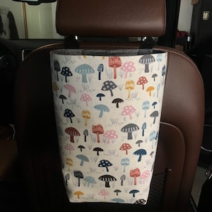May include: A white fabric car seat organizer with a mushroom pattern in shades of blue, pink, yellow, and brown. The organizer is hanging from the headrest of a car seat.