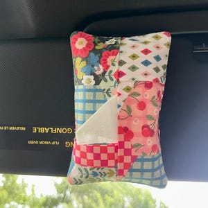 May include: A patchwork tissue holder with a variety of floral, cherry, and geometric patterns in red, pink, blue, and white. The holder is hanging from a car visor.