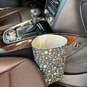 May include: A black and green fabric trash can with a floral pattern hangs from the side of a car seat. The trash can is made of fabric and has a drawstring closure.