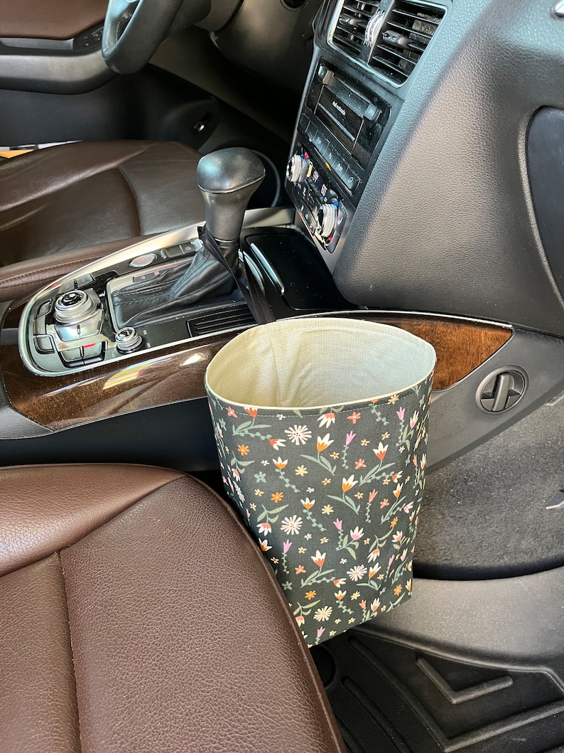 May include: A black and green fabric trash can with a floral pattern hangs from the side of a car seat. The trash can is made of fabric and has a drawstring closure.