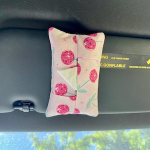 May include: A pink fabric tissue holder with a cherry pattern, attached to a car visor. The holder is rectangular with a slit for tissue dispensing. The cherry design features red cherries with green stems on a light pink background.
