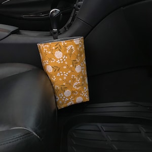 K&ouml;nnte beinhalten: Ein gelber Stoff-M&uuml;llbeh&auml;lter f&uuml;r Autos mit einem wei&szlig;en Blumenmuster, der an der Seite eines Autositzes h&auml;ngt.