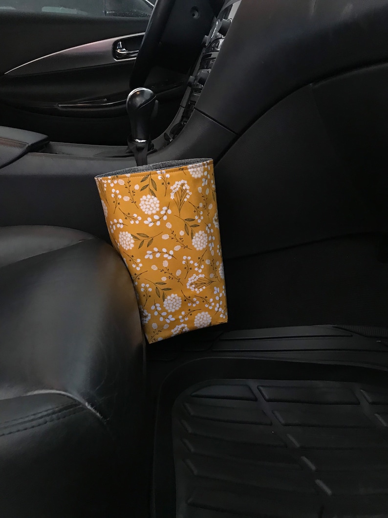 K&ouml;nnte beinhalten: Ein gelber Stoff-M&uuml;llbeh&auml;lter f&uuml;r Autos mit einem wei&szlig;en Blumenmuster, der an der Seite eines Autositzes h&auml;ngt.