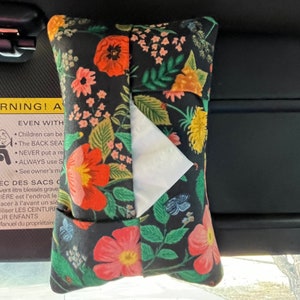 May include: A black and floral patterned tissue box holder with a white tissue box inside. The holder is hanging from a car's sun visor. The text on the holder reads "WARNING! A EVEN WITH Children can be The BACK SEAT NEVER put a re ALWAYS use S See owner's m EC DES SACS C vent être blessés grave IERE est l'endroit le acer à l'avant un siè iliser LES CEINTURE JR ENFANTS Manuel du propriétaire"