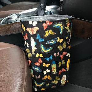 May include: A black fabric car trash can with a butterfly print. The trash can is hanging on the side of a car seat.