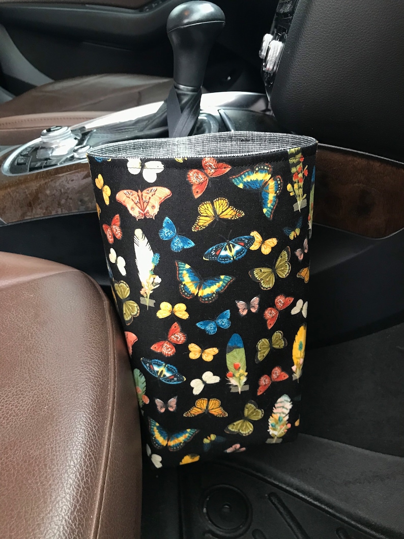 May include: A black fabric car trash can with a butterfly print. The trash can is hanging on the side of a car seat.