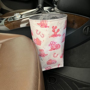 May include: A white fabric car trash bag with a pink cowboy-themed pattern. The pattern includes cowboy hats, cacti, horseshoes, and stars. The bag is cylindrical and hangs from the car's console.