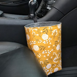 K&ouml;nnte beinhalten: Ein gelber Auto-M&uuml;lleimer mit einem wei&szlig;en Blumenmuster. Der M&uuml;lleimer h&auml;ngt am Schalthebel in einem Auto.