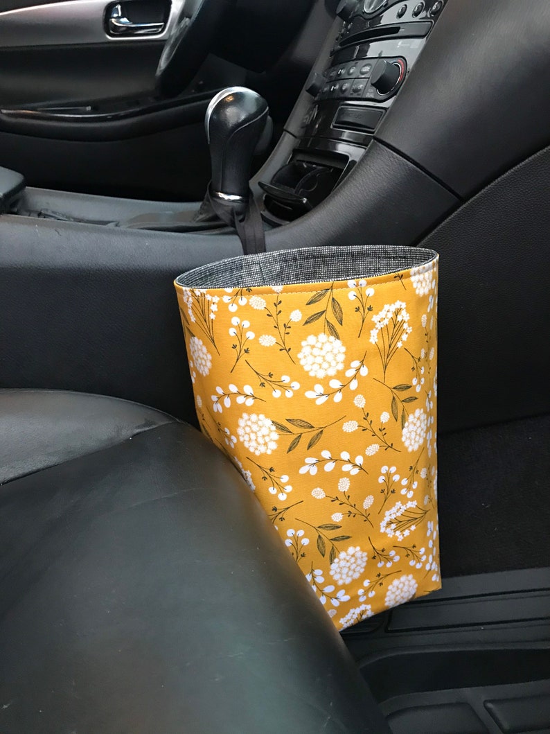 K&ouml;nnte beinhalten: Ein gelber Auto-M&uuml;lleimer mit einem wei&szlig;en Blumenmuster. Der M&uuml;lleimer h&auml;ngt am Schalthebel in einem Auto.