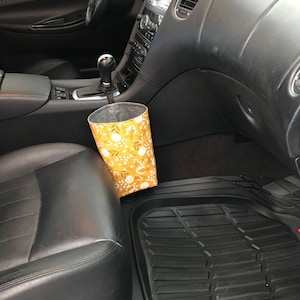 K&ouml;nnte beinhalten: Ein gelber und wei&szlig;er Stoffm&uuml;lleimer mit floralem Muster und einem schwarzen Kordelzug, der oben h&auml;ngt, platziert auf dem Beifahrersitz eines Autos. Das Auto hat schwarze Ledersitze und eine schwarze Fu&szlig;matte.