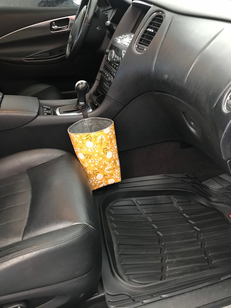 K&ouml;nnte beinhalten: Ein gelber und wei&szlig;er Stoffm&uuml;lleimer mit floralem Muster und einem schwarzen Kordelzug, der oben h&auml;ngt, platziert auf dem Beifahrersitz eines Autos. Das Auto hat schwarze Ledersitze und eine schwarze Fu&szlig;matte.