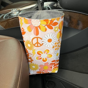 May include: A pink and orange car trash can with a floral and peace sign pattern. The trash can is hanging on the side of a car seat.