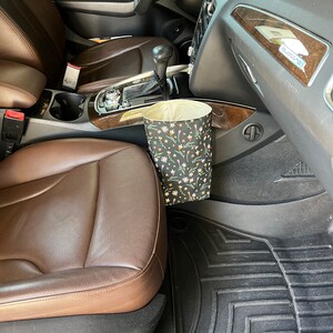 May include: A brown leather car seat with a black and white floral patterned trash can hanging from the side. The car has black floor mats with the WeatherTech logo.
