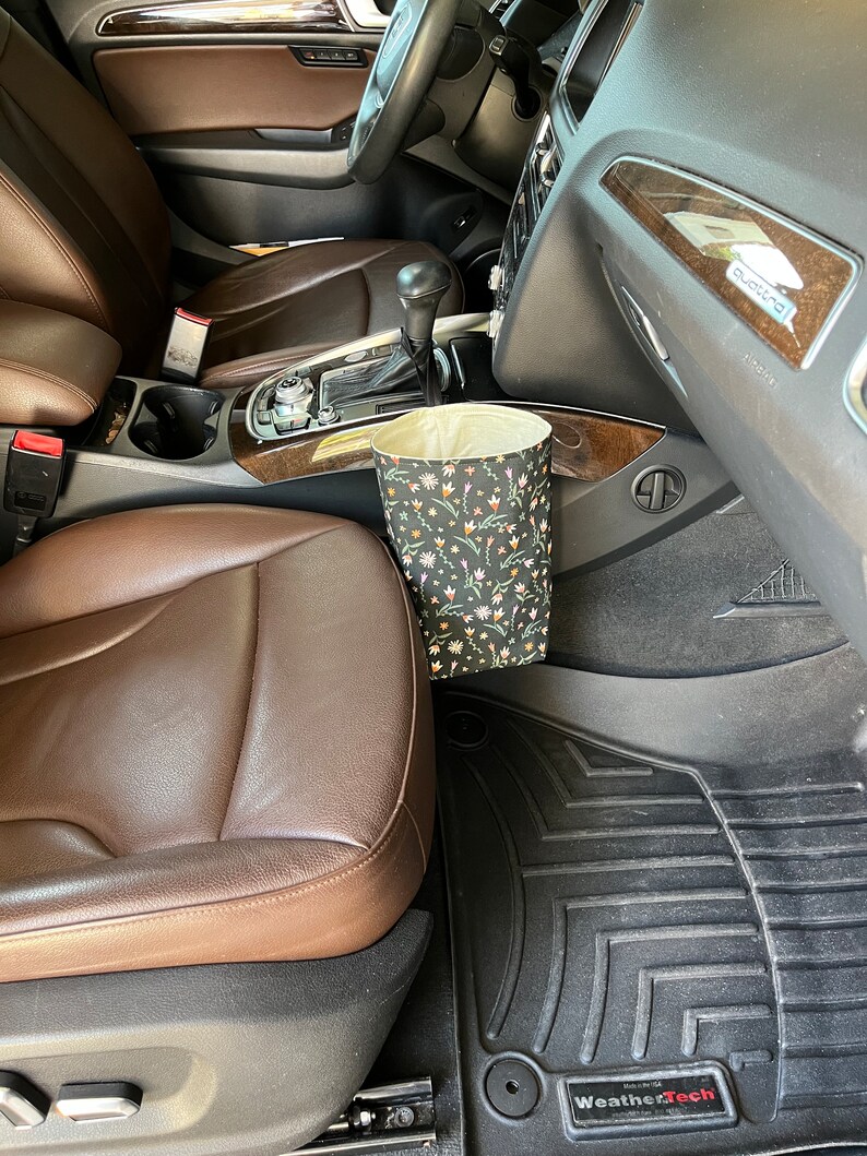 May include: A brown leather car seat with a black and white floral patterned trash can hanging from the side. The car has black floor mats with the WeatherTech logo.