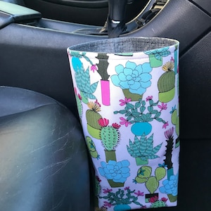 May include: A white fabric car trash can with a cactus and succulent print. The trash can is hanging from the side of a car seat.