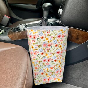 May include: A floral print car trash bag. The bag features a white background with a pattern of pink, yellow, and red flowers, along with blue leaves. The bag is made of fabric and has a gray interior lining.