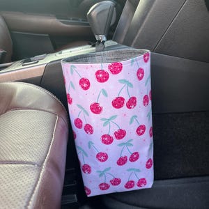 May include: A white fabric car trash can with a cherry print. The trash can is hanging on the side of a car seat.