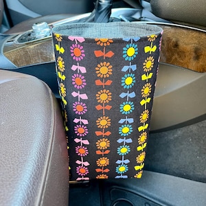 May include: A black fabric car trash bag with a colorful floral pattern. The flowers are in rows of pink, orange, yellow, and blue. The bag is placed in a car's center console.