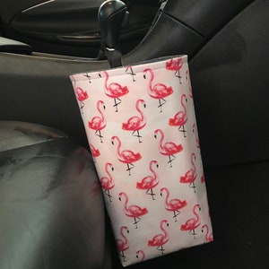 May include: A white fabric car trash can with a pink flamingo pattern. The trash can is hanging on the side of a car seat.