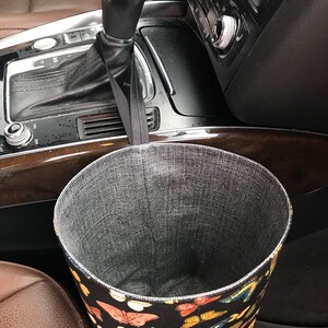 May include: A black fabric trash can with a butterfly print hangs from the gear shift in a car. The trash can is lined with a gray mesh material.