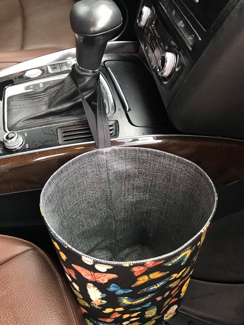 May include: A black fabric trash can with a butterfly print hangs from the gear shift in a car. The trash can is lined with a gray mesh material.