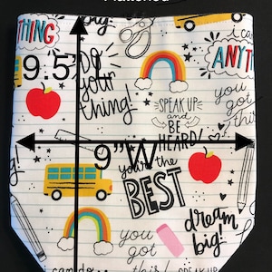 K&ouml;nnte beinhalten: Eine wei&szlig;e Stofftasche mit einem Schul-Motiv. Die Tasche zeigt einen gelben Schulbus, rote &Auml;pfel, Regenb&ouml;gen und inspirierende Spr&uuml;che wie "You got this!" und "Dream big!" Die Tasche ist 24,1 cm hoch und 22,9 cm breit.