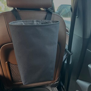 May include: A black fabric trash can hangs from the headrest of a car seat. The can is made of a durable material and has a wide opening for easy access.
