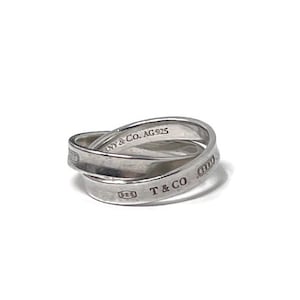 May include: A silver ring with an overlapping design. The ring is engraved with "NY & Co. AG925" and "T & CO". The ring is made of a shiny, reflective metal.