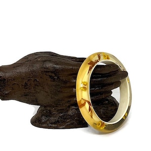May include: A clear resin bangle bracelet with a gold-toned finish and embedded dried flowers. The flowers are a mix of brown and yellow.