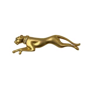 May include: A gold-toned brooch shaped like a leaping panther in a stretched, running pose. The panther features a smooth, matte finish and wears a collar adorned with a row of small, clear, sparkling stones around its neck.