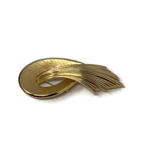 May include: A gold-toned abstract brooch with a curved shape and a flowing design. The brooch has a textured surface and a long, thin, flowing tail.