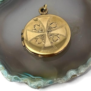 May include: A round gold-toned metal locket featuring an engraved cross design with floral motifs at the center. The locket has a small circular bail at the top for attachment and a visible hinge on the left side, showcasing a polished metallic finish.